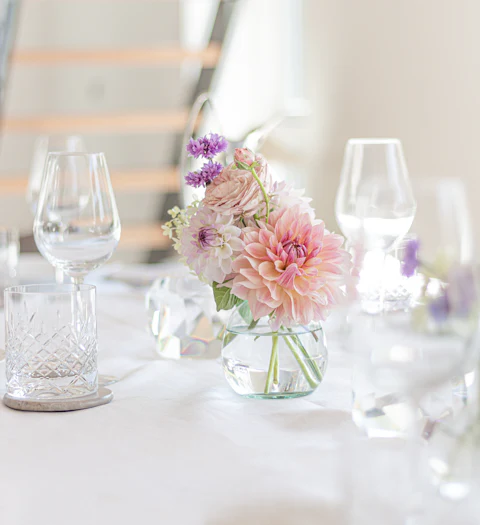 Small glass vase of dahlias and lavender on a white dinner table set with wine glasses